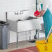 A man in a blue uniform using a yellow mop to clean a Regency commercial utility sink.