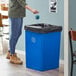 A woman pouring water from a plastic bottle into a blue Lavex square recycle bin.