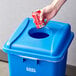 A hand throwing a can of soda into a blue Lavex recycle bin with a bottle/can hole.