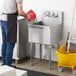 A person wearing a black apron pouring water into a Regency stainless steel utility sink on a counter in a professional kitchen.