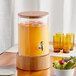 An Acopa glass beverage dispenser with orange juice and a wooden lid on a table.