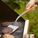 A person using an Outset stainless steel turner to cook meat on a grill.