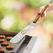 A hand holding an Outset stainless steel slotted turner with a bamboo handle over a grill.