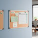 A 24 by 18 inch wall-mount cork board with an aluminum frame, displayed with various notes and a calendar.