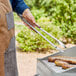 A person using Outset stainless steel tongs with rosewood handles to cook meat on a grill.