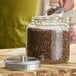 A large clear glass jar filled with coffee beans, shown with a brushed metal lid resting beside it.