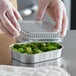 A gloved hand holding a clear container of broccoli.