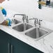 A person washing dishes in a Waterloo stainless steel three compartment drop-in sink with a swing faucet.