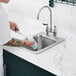 A person with tattoos on their arm using a toothbrush to clean a glass in a Waterloo stainless steel drop-in sink.