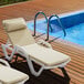 Two Lancaster Table & Seating white resin chaise lounges with cream cushions and pillows next to a pool.