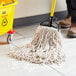 A person in a brown boot cleaning a floor with a yellow handled Lavex Natural Cotton Wet Mop.