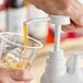 A hand using a Tablecraft syrup pump to fill a plastic cup with liquid.