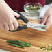 A pair of hands using Mercer Culinary stainless steel kitchen shears to cut green onions on a cutting board.