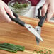 A person using Mercer Culinary stainless steel kitchen shears with black handles to cut chives on a cutting board.
