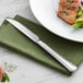 An Acopa Landsdale stainless steel dinner knife on a napkin next to a plate of salmon and vegetables.