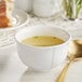 A white Acopa porcelain bouillon cup filled with soup and a spoon on a table with bread.