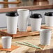 A group of white Choice paper hot cups with black lids on a wooden tray.