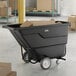 A black forkliftable tilt truck or trash cart with a 1 cubic yard capacity, shown holding cardboard boxes in a warehouse setting.