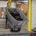 A woman in a yellow vest pushing a Lavex grey heavy-duty tilt truck full of garbage bags.