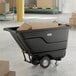 A black heavy-duty tilt truck or trash cart with a 0.5 cubic yard capacity, shown holding cardboard boxes in a warehouse setting.