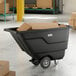 A black Lavex tilt truck or trash cart with a 0.5 cubic yard capacity, shown holding cardboard boxes in a warehouse setting.