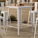 A white Lancaster Table & Seating bar height table on an outdoor patio with a drink on it.