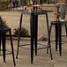 A Lancaster Table & Seating onyx black bar height table with black chairs on a stone patio.