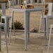 A Lancaster Table & Seating nickel gray bar height outdoor table with drinks on it and chairs on a patio.