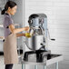 A woman in an apron pouring liquid from a measuring cup into a Centerline commercial stand mixer.