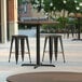 A Lancaster Table & Seating Excalibur round counter height table with a textured brown surface and cross base plate, outside on a patio with three stools.