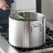 A hand holding a black-handled spoon stirring broccoli in a Choice aluminum sauce pot.