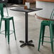 A square bar height table with a smooth black finish and a cross base plate.