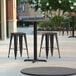 A Lancaster Table & Seating round counter height table with a cross base and three black stools on a patio.