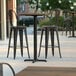 A Lancaster Table & Seating bar height table with a textured farmhouse finish and cross base plate on a patio with black metal bar stools.