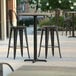 A Lancaster Table & Seating bar height table with a textured canyon metal finish and cross base plate on a patio with chairs.