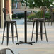 A Lancaster Table & Seating Excalibur bar height table with a textured metal finish and cross base plate with two black chairs on a patio.