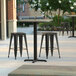 A Lancaster Table & Seating square counter height table with a textured metal finish and cross base plate on a patio with two black stools.