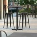 A Lancaster Table & Seating bar height table with a cross base on an outdoor patio with three black chairs.