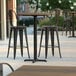 A Lancaster Table & Seating bar height table with a textured farmhouse finish and cross base plate on an outdoor patio with chairs.