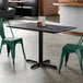 A rectangular standard height table with a smooth dark finish and a black cross base plate, shown with green metal chairs.