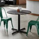 A round black table with a smooth finish and cross base plate, accompanied by two green metal chairs.
