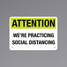 A black and yellow rectangular sign on a wall that says "Attention / We're Practicing Social Distancing"