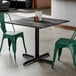 A square standard height table with a smooth dark finish and a black cross base plate, accompanied by two green metal chairs.