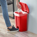 A person using a red Lavex step-on trash can to throw a white bag away.