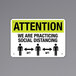 A black and yellow aluminum sign with the text "Attention / We Are Practicing Social Distancing" and people icons.