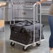 A woman using a Vesture aluminum cart to carry a black delivery bag in a kitchen.