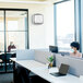 A woman wearing a mask sits at a white table using a laptop with a white AeraMax 9416201 PRO III Wall-Mount Air Purifier on the wall.