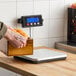 A low-profile digital receiving scale with a remote display mounted on a wall, shown being used to weigh a wooden box of oranges.