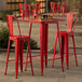 A Lancaster Table & Seating ruby red metal bar height table with two ruby red chairs on an outdoor patio.