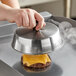 A hand using an American Metalcraft stainless steel basting cover to melt cheese on a burger.